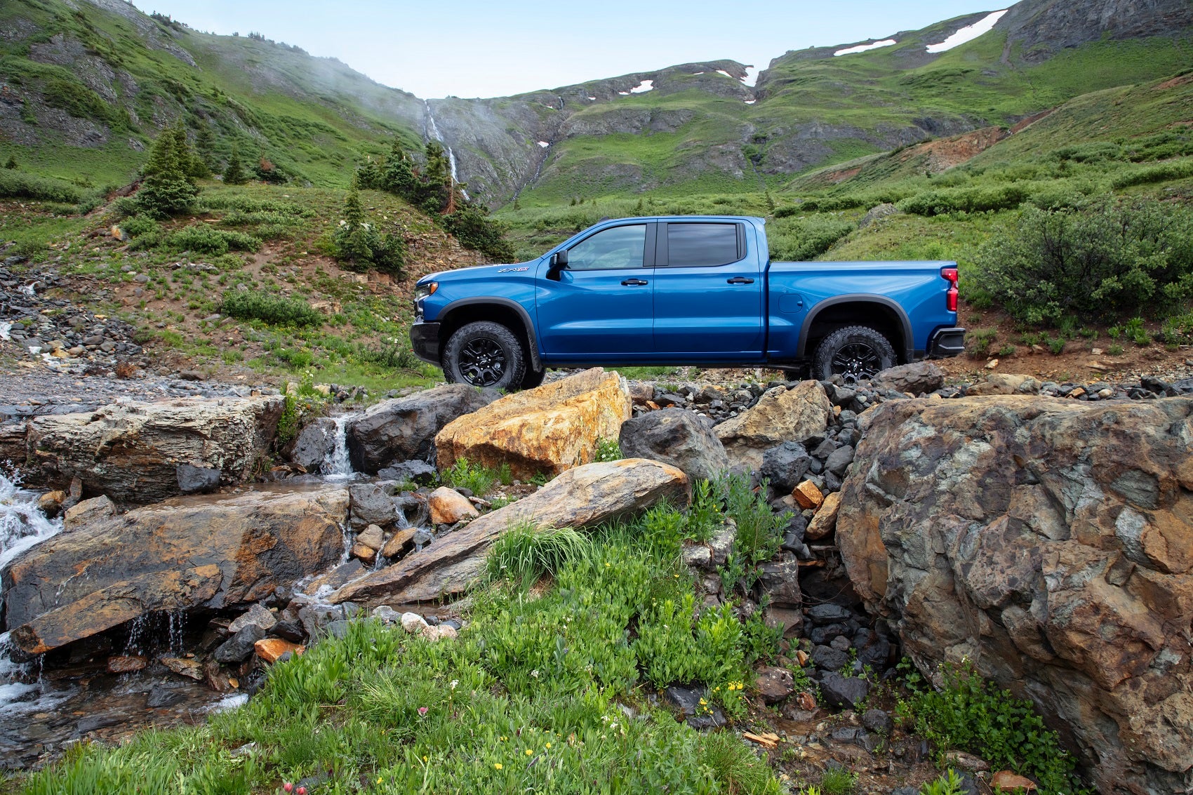 blue chevy truck drives over rocks with scenic mountain view in background